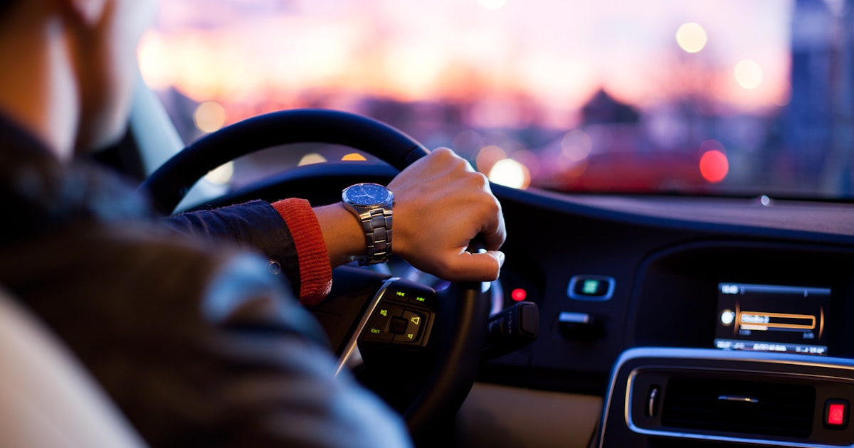 Driver holding a steering wheel at dusk with blurred city lights ahead